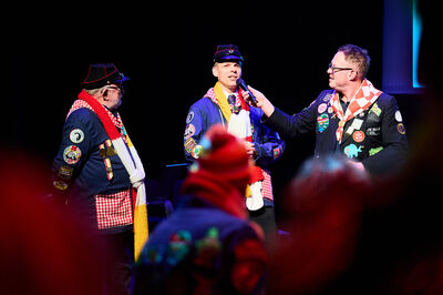Drie mannen in kleurrijke scoutsuniformen staan op een podium; één vertelt iets in een microfoon.