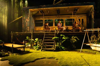 Een theatrale scène toont een opengezaagde treinwagon die midden in een bos staat. Binnen zitten vier personen op busstoelen, terwijl rondom de wagon een groen, sprookjesachtig decor met planten, een steiger, een hangmat en een schommel te zien is. Warm licht verlicht het geheel, waardoor het lijkt alsof de wagon deel uitmaakt van het bos.