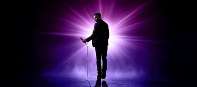 Een sfeervol portret van cabaretier Rayen Panday, van achteren gefotografeerd tegen een felpaarse lichtbron die als een ster uiteenvalt in lichtstralen. Hij draagt een donker pak, houdt een microfoon met draad vast en kijkt over zijn schouder achterom naar de camera. De voorgrond is donker met een lichte mistlaag over de vloer.