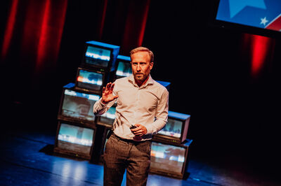 Raymond Mens staat in een wit overhemd en een donkere broek op een verlicht podium. Hij maakt een gebaar met zijn rechterhand terwijl hij een presentatie geeft. Achter hem staat een opstapeling van oude televisies die verschillende beelden tonen tegen een achtergrond van sfeervolle rode gordijnen.