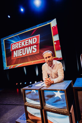 Een close-up van Raymond Mens die over een stapel oude monitoren leunt op een podium. Hij heeft zijn vingers in elkaar geslagen en kijkt serieus in de camera. Direct achter hem hangt een groot scherm met de tekst 'BREKEND NIEUWS' in een rood kader. De monitoren op de voorgrond tonen beelden van de Amerikaanse vlag.