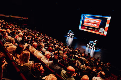 Een volle theaterzaal kijkt vanaf de zijkant naar een verlicht podium. Op het grote scherm staat de tekst 'WIJ WACHTEN OP VERBINDING...' tegen een rood-blauwe achtergrond met sterren. Onder de tekst staat een oproep om naar een podcast te luisteren met een QR-code. Op het podium staan twee stapels oude televisies die beelden vertonen.