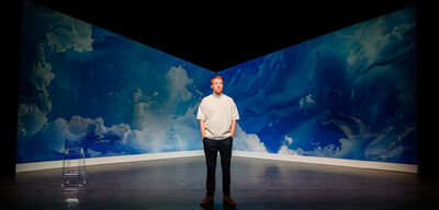 Rene van Meurs haar staat alleen midden op een theaterpodium met een blauwe, wolkachtige achtergrond; zijn handen zitten in zijn zakken en links van hem staat een transparante kruk.