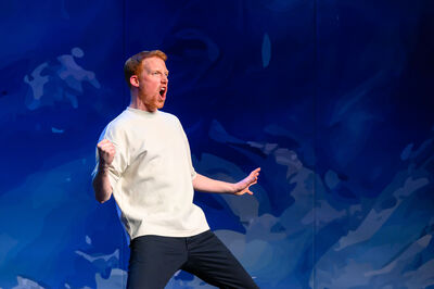 Rene van Meurs staat op een podium tegen een blauwe achtergrond, leunt achterover met gebalde vuist en open mond alsof hij roept, in een energieke en expressieve pose onder theaterbelichting.