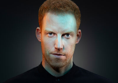 Close-up van Rene van Meurs met kort rood haar en lichte baard die recht in de camera kijkt met een intense, serieuze blik tegen een donkere achtergrond met subtiele belichting.