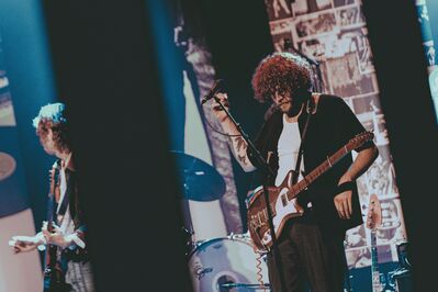 Alt-tekst: Een gitarist met krullend haar staat op een podium naast een microfoonstandaard en houdt zijn elektrische gitaar vast. Op de achtergrond zijn een drummer en een tweede gitarist te zien. De scène is verlicht met koel blauw licht en heeft een intieme livesfeer.