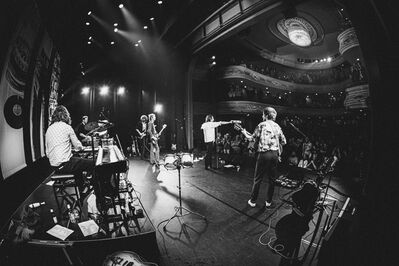 Zwart-witfoto van een band die optreedt in een theaterzaal. De muzikanten staan op het podium met gitaren, drums en een toetseninstrument, terwijl het publiek zichtbaar is op de vloer en op de balkons. Spotlights verlichten het podium en een grote kroonluchter hangt aan het plafond, wat zorgt voor een klassieke concertsetting.