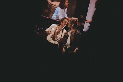 Twee muzikanten staan dicht bij elkaar op het podium en zingen samen in één microfoon. Eén van hen houdt een tamboerijn vast, terwijl de ander een gitaar draagt. Op de achtergrond speelt een toetsenist. De foto is genomen vanuit de coulissen, waardoor de scène wordt omlijst door donkere randen en een intieme concert­sfeer ontstaat.