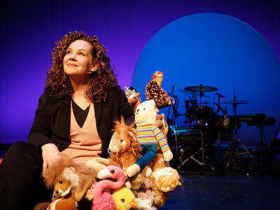 Sara Kroos zit op een podium en kijkt glimlachend omhoog, omringd door een stapel knuffeldieren; op de achtergrond zijn een drumstel en een blauw-paarse theaterbelichting zichtbaar.