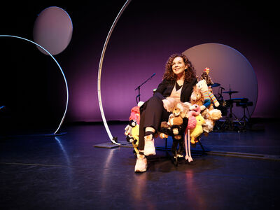 Sara Kroos zit glimlachend in een stoel die bedekt is met knuffeldieren op een theaterpodium met paarsblauwe belichting; op de achtergrond staan een drumstel en grote ronde decorvormen.