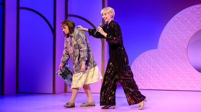 Alt-tekst: Twee Bianca in alledaagse kleding met een plastic tas loopt voorovergebogen, terwijl een glimlachende Plien in een glinsterende zwarte outfit met microfoon haar speels vooruit duwt tegen een paars verlichte achtergrond.