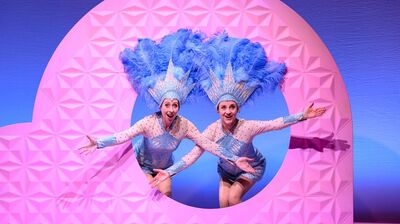 Plien & Bianca in sprankelende blauwe showkostuums met grote verenhoeden steken lachend hun armen uit door een ronde opening in een roze decor.