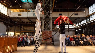 In een industriële hal voeren vier performers een acrobatische scène uit voor een zittend publiek. Twee personen tillen elk een andere performer omhoog: links staat iemand rechtop op de schouders van een ander, rechts wordt een persoon in een rode outfit horizontaal boven het hoofd gedragen. Het publiek zit in de achtergrond van de speelvloer en kijkt aandachtig toe.