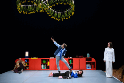 Theatrale scène op een podium met zwarte achtergrond en een lange felrode kast. Boven het podium hangen slingers van gele tulpen. In het midden staat een man in overhemd en stropdas met één voet op een vrouw die op de grond ligt; hij houdt zijn telefoon omhoog alsof hij een selfie maakt. Links zit een andere man tegen de kast op de grond en rechts staat een vrouw in witte kleding die toekijkt. De scène oogt absurd en dramatisch.
