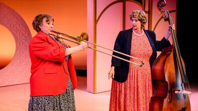 Plein & Bianca op een kleurrijk theaterpodium: links speelt Bianca in een rode blazer trombone, terwijl rechts Plien in een lange rode jurk en donker jasje een contrabas vasthoudt en verbaasd naar haar kijkt.