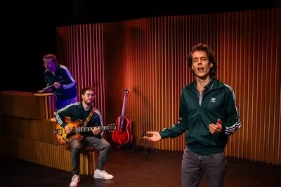 Drie mannen staan op een verlicht podium voor een wand van verticaal geplaatst bruin karton. De voorste man zingt met een open gebaar, terwijl de tweede man op de achtergrond zittend op een elektrische gitaar speelt. Een derde man staat linksachter en bespeelt een percussie-instrument van karton. Alle drie dragen ze donkergroene Adidas-trainingsjacks met witte strepen.