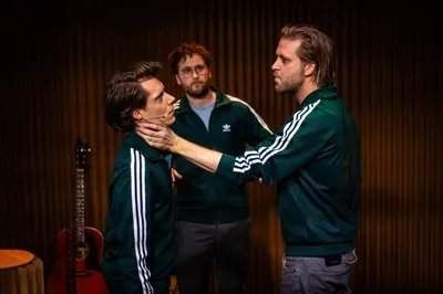 Drie mannen in donkergroene Adidas-trainingsjacks staan op een podium voor een achterwand van verticaal karton. De rechter man houdt met een serieuze, indringende blik de nek van de linker man vast, die hem gespannen aankijkt. De derde man staat in het midden op de achtergrond en kijkt met een neutrale uitdrukking toe. Links op de achtergrond staat een rode akoestische gitaar.