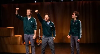 Drie mannen in donkergroene Adidas-trainingsjacks staan op een podium voor een achterwand van verticaal bruin karton. De linker en middelste man heffen krachtig hun rechtervuist in de lucht terwijl ze zingen of roepen. De rechter man kijkt hen met een vragende en licht verbaasde blik aan. Op de achtergrond staat een elektrische gitaar tegen de kartonnen wand.
