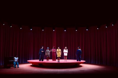 Zes mensen in verschillende outfits staan op een verhoogd podium, omringd door donkere gordijnen en verlichting.