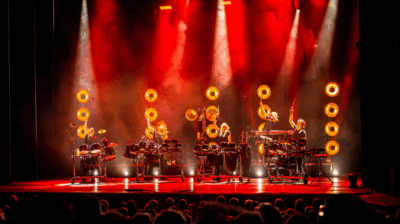 Vier percussionisten spelen op uitgebreide drumopstellingen op een groot podium dat fel rood verlicht is. Achter hen hangen meerdere ronde lampen die warm oranje licht uitstralen. De muzikanten houden hun drumstokken omhoog in een krachtig moment, terwijl onderaan in beeld het publiek zichtbaar is als donkere silhouetten.