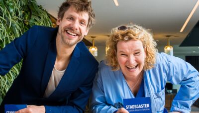 Twee lachende mensen staan binnen en houden een boek in hun handen, met een groene muur op de achtergrond.