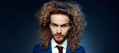 Teun van den Elzen met lange, volle krullen en een haarband kijkt serieus in de camera; hij draagt een blauw pak met wit overhemd en rode stropdas tegen een donkere achtergrond.