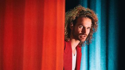 Teun van den Elzen met lange krullen en een rode blazer kijkt vanachter een rood theatergordijn de zaal in; op de achtergrond hangen blauwgroene gordijnen.