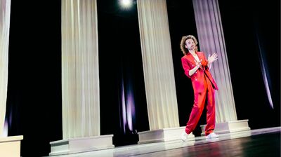 Teun van den Elzen met lange krullen in een felrood pak staat op een theaterpodium tussen hoge witte zuilen en maakt een handgebaar alsof hij iets uitlegt.