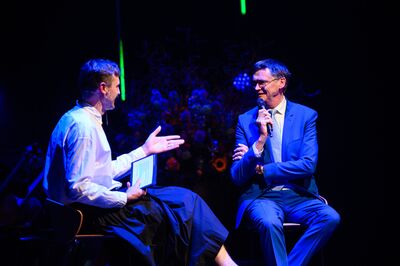 Twee mannen in zakelijke kleding voeren een gesprek op een podium, omringd door kleurrijke lichten en bloemen.