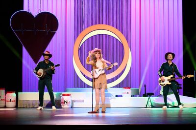 Drie muzikanten staan op een kleurrijk verlicht theaterpodium. In het midden staat Tineke Schouten met een gitaar en een roze cowboyhoed bij een microfoon. Links en rechts van haar staan twee muzikanten met snaarinstrumenten. Op de achtergrond hangen paarse gordijnen, een grote verlichte cirkel en een hartvorm met een lichtlijn.