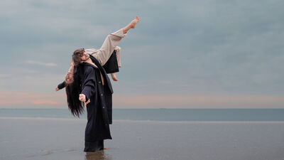 Twee personen staan in ondiep water bij de zee tijdens schemering. Eén persoon tilt de ander achterover in een sierlijke, dansachtige houding, terwijl de horizon en een bewolkte lucht op de achtergrond zichtbaar zijn.