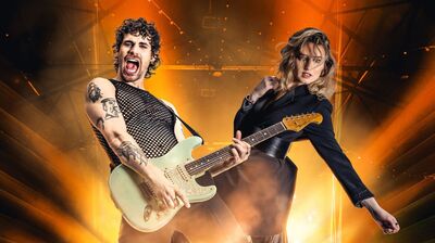 Twee muzikanten poseren op een podium met fel oranje licht en rook op de achtergrond. Links staat een gitarist met krullend haar en zichtbare tatoeages die enthousiast speelt op een elektrische gitaar. Rechts staat een vrouw in een donkere outfit in een krachtige pose. De scène heeft een energieke, rockachtige uitstraling.