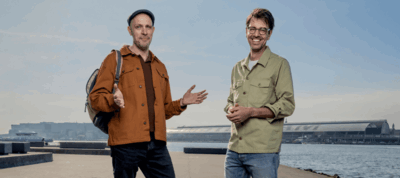 Twee lachende mannen staan buiten op een kade bij het IJ in Amsterdam, met op de achtergrond het Centraal Station onder een heldere, blauwe lucht. De linker man draagt een bruine jas, een rugzak en een pet, en maakt een verwelkomend gebaar met zijn handen. De rechter man draagt een groen overhemd en een bril.