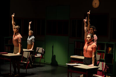 Vier personen staan achter schoolbanken in een donker verlicht klaslokaal en steken hun rechterarm recht omhoog met één vinger omhoog. Op de achtergrond zijn boekenkasten en een klok aan de muur te zien. Het toneel is gericht belicht, waardoor sterke schaduwen op de vloer en tegen de achterwand ontstaan.
