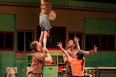 Drie performers ondersteunen een vierde die ondersteboven in een handstand wordt vastgehouden op een theaterpodium met een klaslokaaldecor. Twee personen houden de handen van de acrobaat stevig vast, terwijl een derde met opgeheven armen klaarstaat om te helpen. Op de achtergrond zijn groene kasten, een schoolbord en een klok zichtbaar.