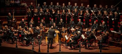 Een groot koor en orkest voeren een klassiek concert uit in een concertzaal. Op de voorgrond dirigeert een dirigent het orkest met strijkers en blazers, terwijl daarachter een koor in zwarte kleding op verhoogde rijen staat te zingen. De musici zitten op een houten podium met rode stoelen en bladmuziekstandaards.