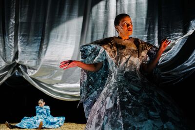 Theatrale scène met dramatische belichting. Op de voorgrond staat een vrouw in een sculpturale jurk met een marmerachtig patroon, haar armen licht gespreid en haar blik omhoog gericht. Achter haar hangt een groot, zilverkleurig doek dat golvend naar beneden valt. Op de achtergrond zit een tweede vrouw in een blauwe jurk op de grond. De sfeer is intens en visueel indrukwekkend.