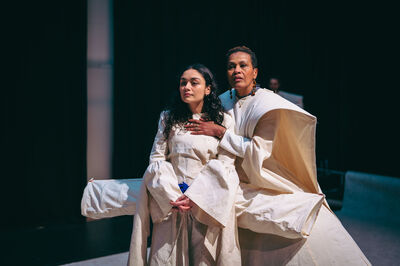 Twee vrouwen in lichte, ruimvallende kostuums zitten dicht bij elkaar op een minimalistisch podium. De oudere vrouw zit achter de jongere en legt beschermend een hand op haar borst, terwijl beiden ernstig voor zich uit kijken. Het decor is sober met donkere gordijnen op de achtergrond en zachte, gerichte belichting die de intieme en intense sfeer benadrukt.