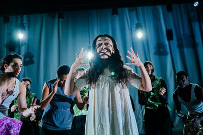 Een acteur in een dramatische houding staat op het podium, omringd door andere performers in een theatrale setting.