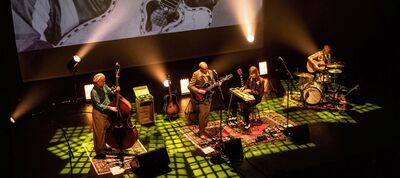 Vier muzikanten spelen samen op een podium dat warm verlicht is met geelgroene spotlights. Links staat een contrabassist, in het midden een gitarist en een muzikant achter een klein toetsinstrument, en rechts een drummer. Ze staan op losse tapijten en achter hen wordt een groot zwart-wit beeld geprojecteerd, wat zorgt voor een intieme, sfeervolle setting.