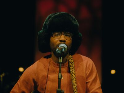 Close-up van een zanger die in een microfoon zingt op een donker podium. Hij draagt een grote, zachte muts, een bril en een warme trui, met een gitaarband over zijn schouder. De achtergrond is onscherp en rood verlicht, wat zorgt voor een intieme en sfeervolle uitstraling.