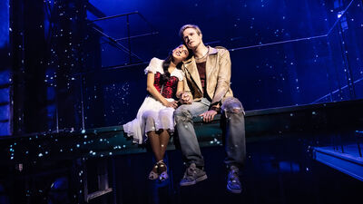 Een jonge vrouw en man zitten naast elkaar op een verhoogd podium in blauw licht, met sterachtige lichtpuntjes op de achtergrond. Ze houden elkaars hand vast en leunen tegen elkaar aan, terwijl ze dromerig omhoog kijken. De sfeer is romantisch en theatraal.