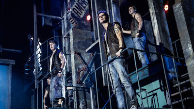 Meerdere mannen staan in het decor van West side story met metalen trappen en loopbruggen. Ze dragen stoere outfits met mouwloze vesten en spijkerbroeken en kijken vastberaden de zaal in. De scène is verlicht met koel blauw licht en heeft een ruige, musicalachtige uitstraling.