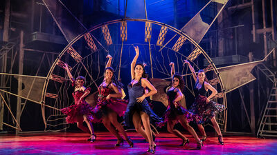 Vijf danseressen voeren een energieke choreografie uit op een theaterpodium. Ze dragen kleurrijke jurken met zwierige rokken en hoge hakken. Achter hen staat een grote metalen constructie in de vorm van een klok met Romeinse cijfers, terwijl de industriële achtergrond en warme belichting zorgen voor een dynamische, musicalachtige sfeer.